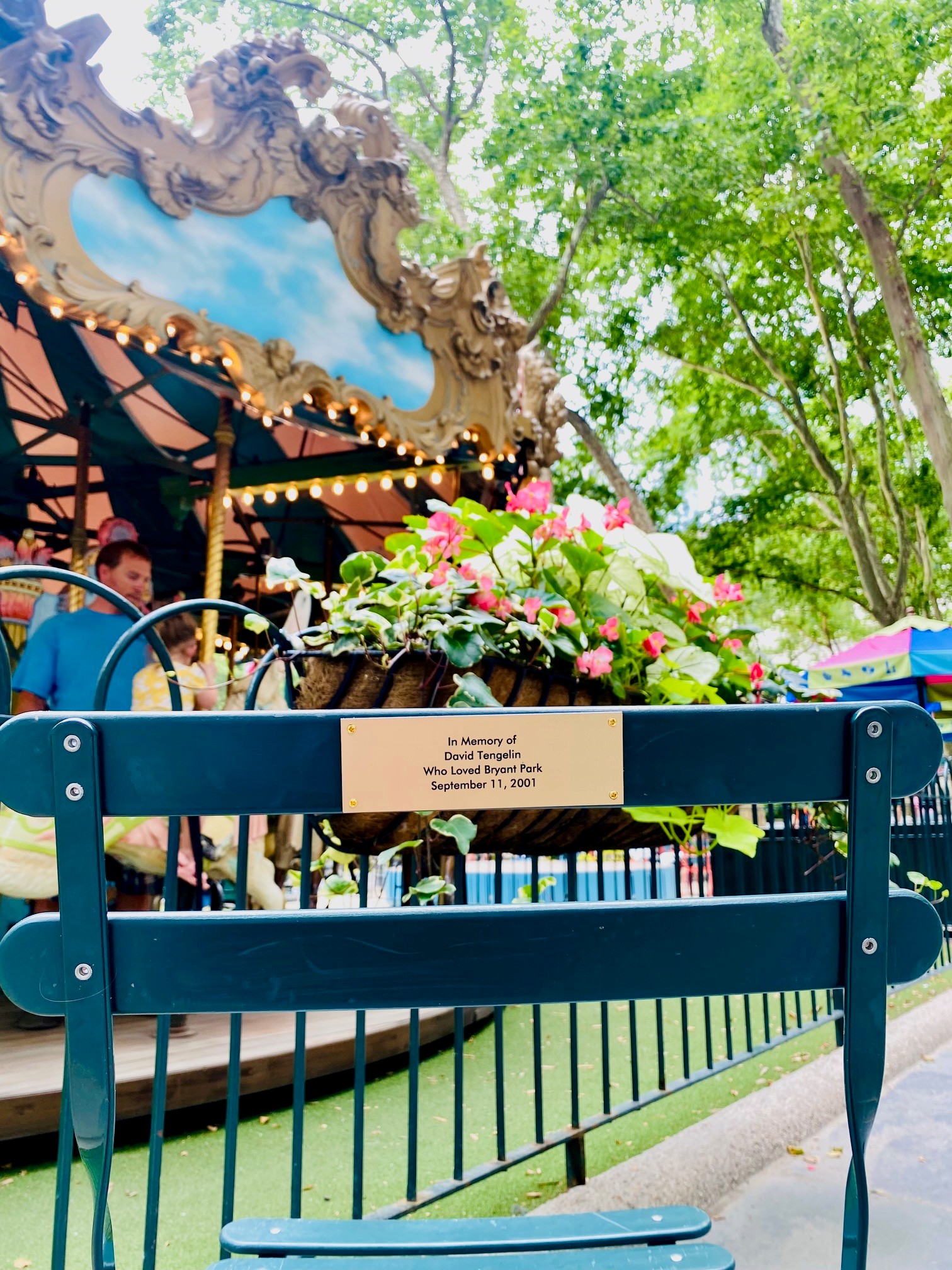 Close view of the memorial plaque on a Bryant Park chair with the carousel, flowers, trees, and a parent and child visible in the background.