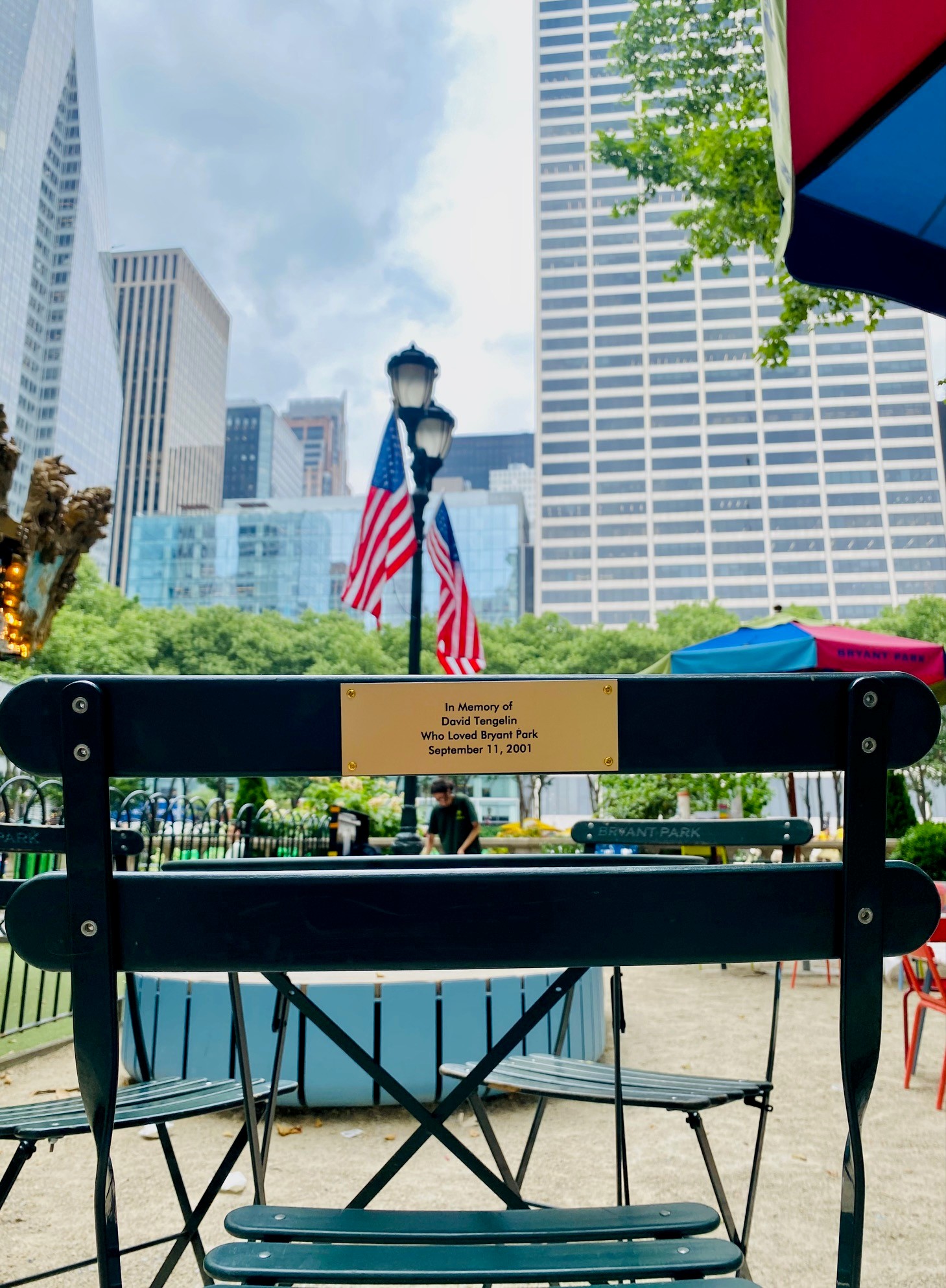 Memorial chair in Bryant Park with American flags, summer trees, surrounding buildings, and visitors enjoying the park in the background.