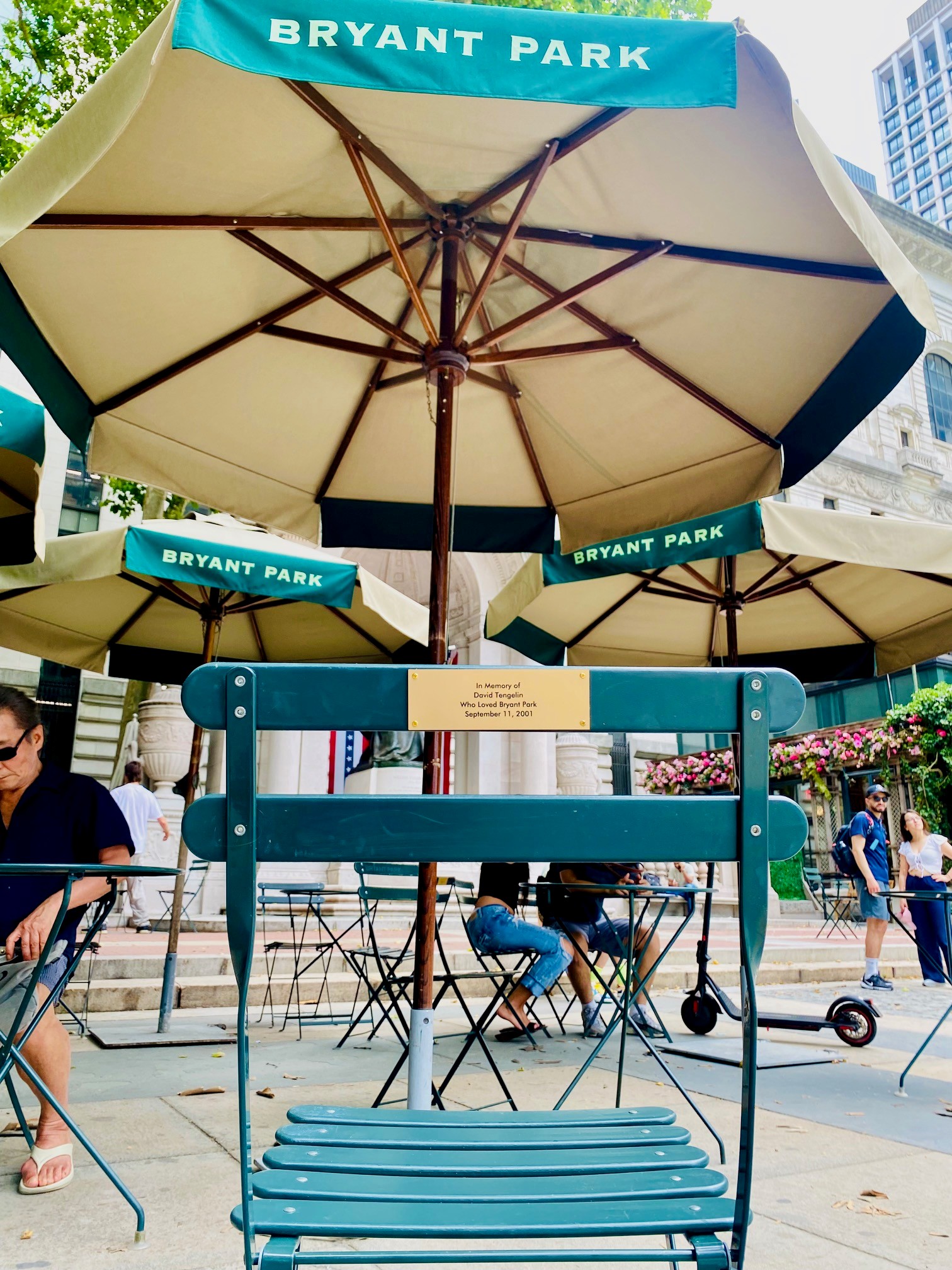 Memorial chair bearing David Tengelin’s plaque beneath Bryant Park umbrellas, surrounded by visitors enjoying the park on a summer afternoon.